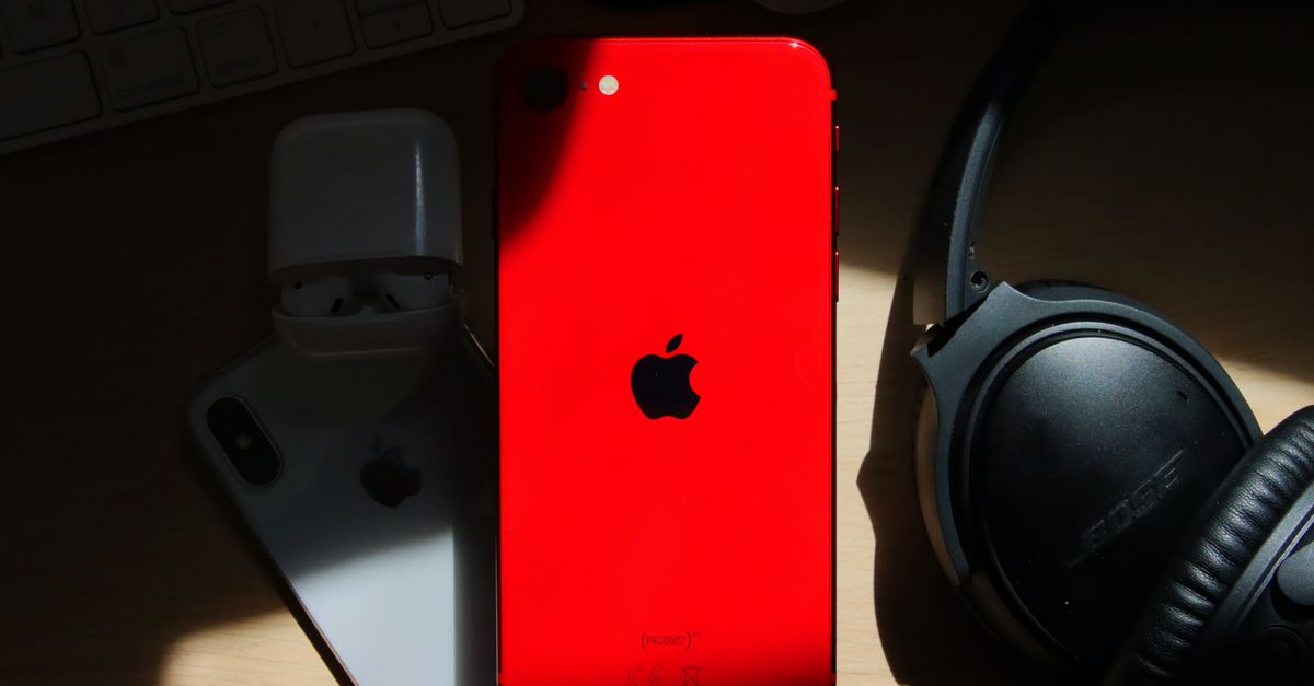 A flat lay of a red iPhone surrounded by AirPods and Bose headphones on a wooden surface.